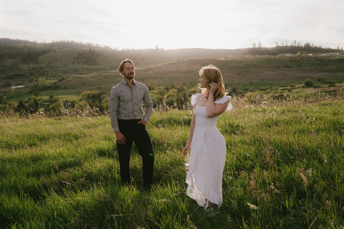 Engaged couple standing apart in a green spring meadow above the Okanagan Valley, the sun low over the hills