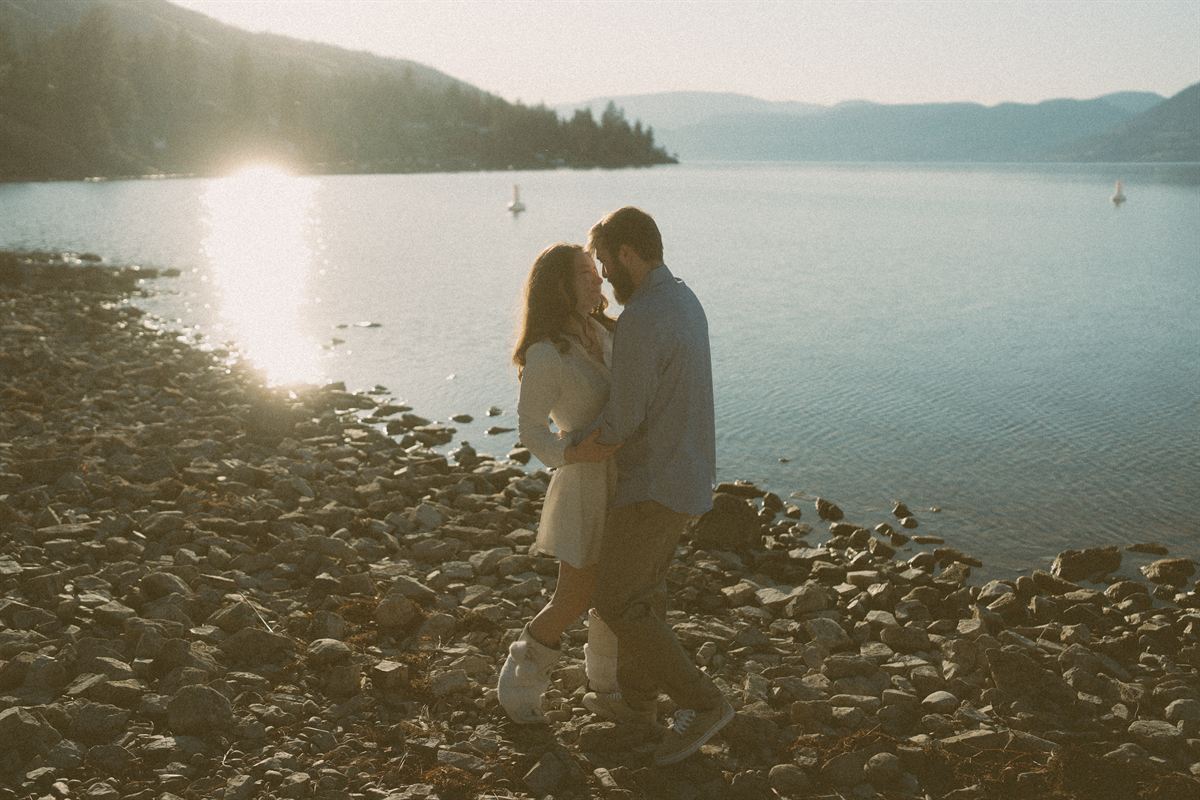 Couple walking the stony shoreline of Okanagan Lake at golden hour, the hills folding away into the distance
