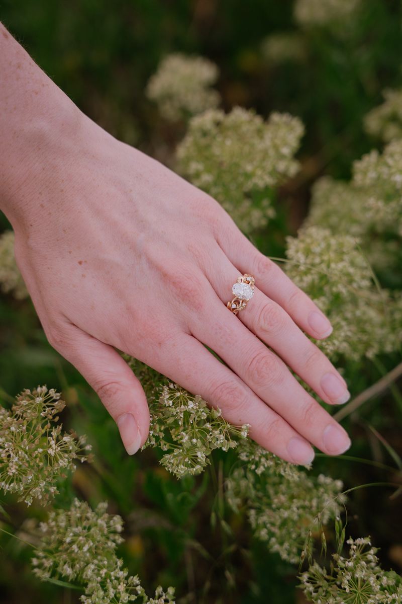 Her hand resting on wild yarrow, an oval engagement ring in a gold setting