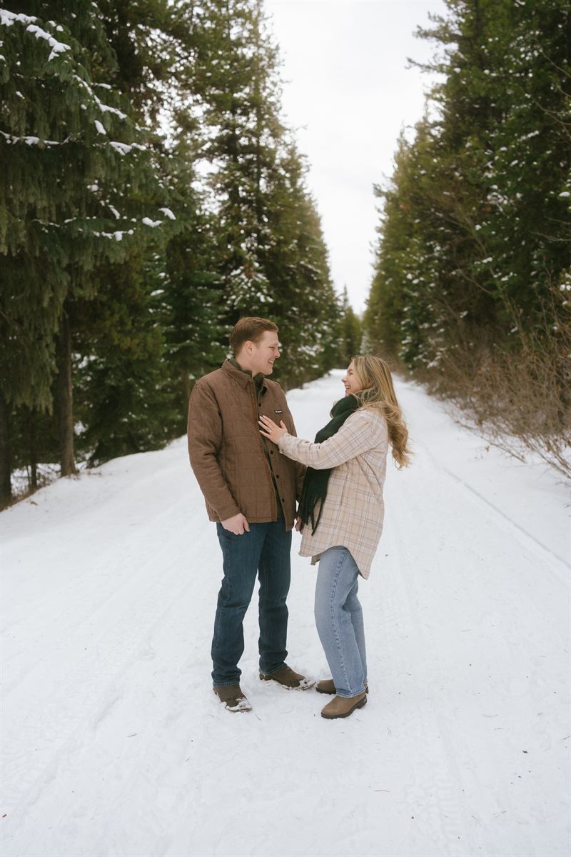 Couple laughing on a snow-covered road between rows of evergreens during a winter engagement session