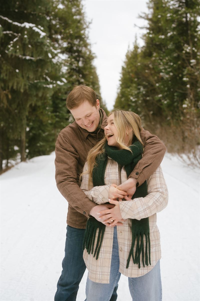 He wraps his arms around her on a snowy forest road, both of them laughing, her engagement ring catching the light