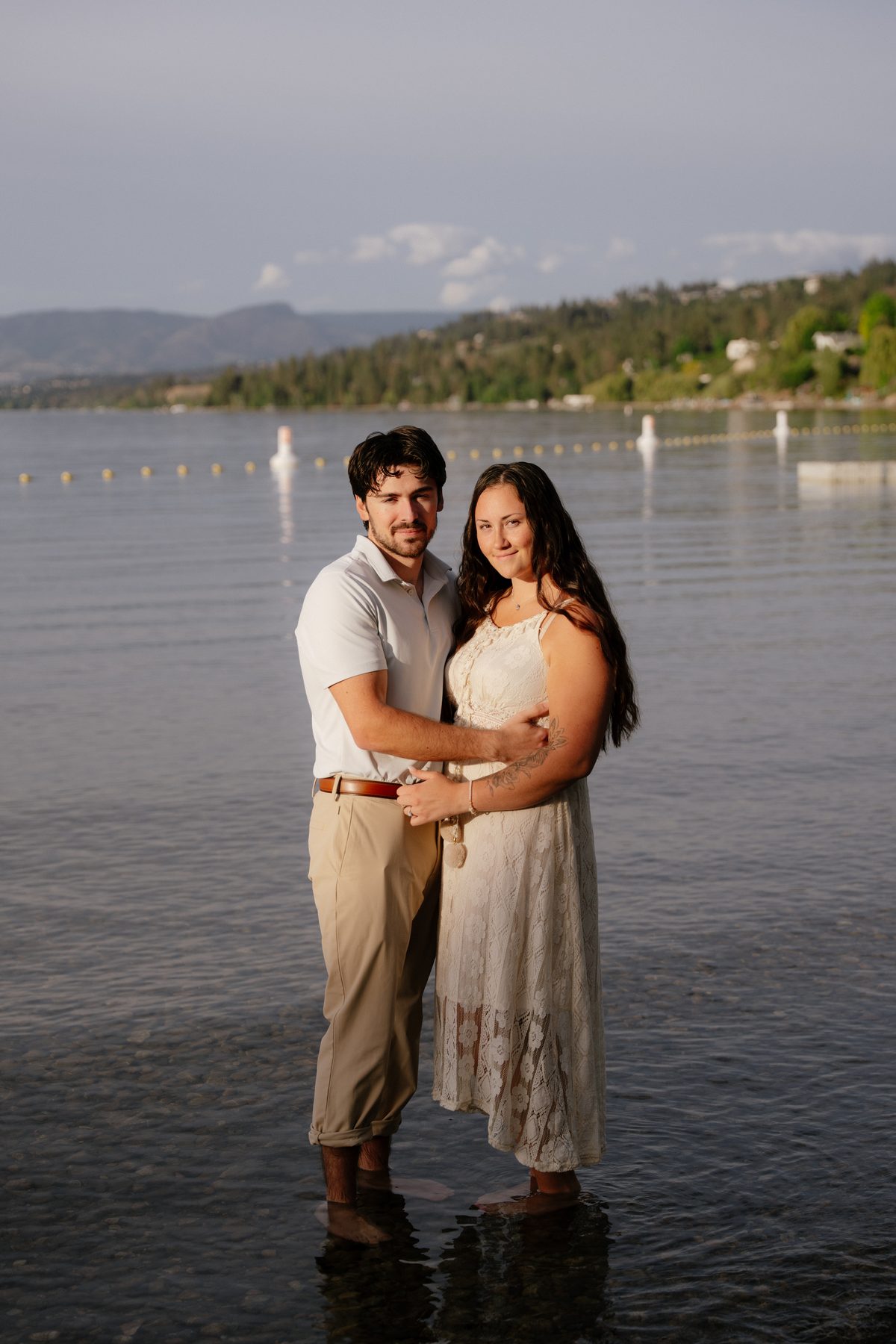 Couple standing ankle-deep in Okanagan Lake, the benchland and hills of the valley behind them