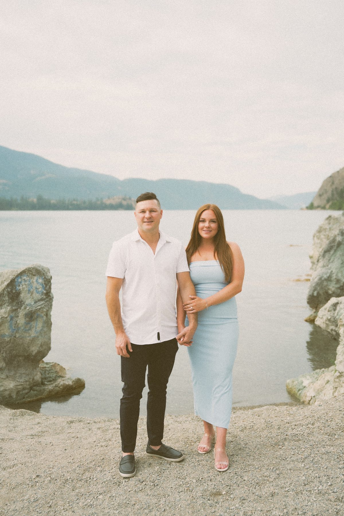 Engaged couple standing arm in arm on the shore of Okanagan Lake under a soft overcast sky