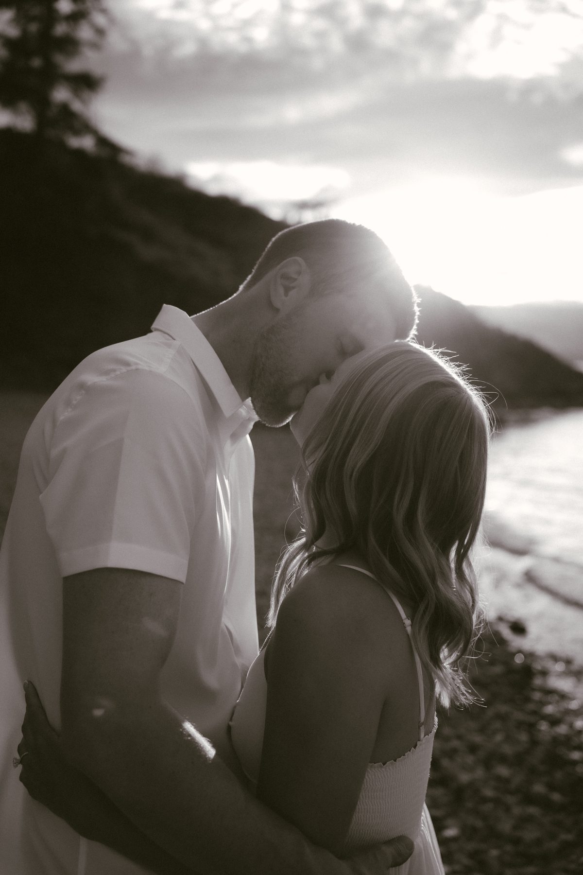 Black and white photo of a couple on the shore of Okanagan Lake at sunset, his forehead resting against hers as the sun flares behind them