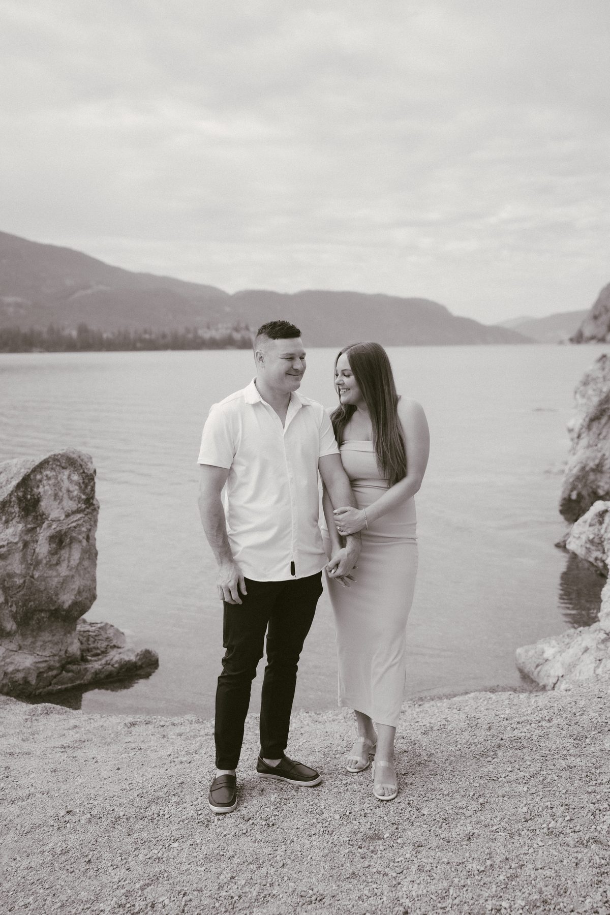 Black and white photo of a couple walking arm in arm along the lakeshore, laughing together
