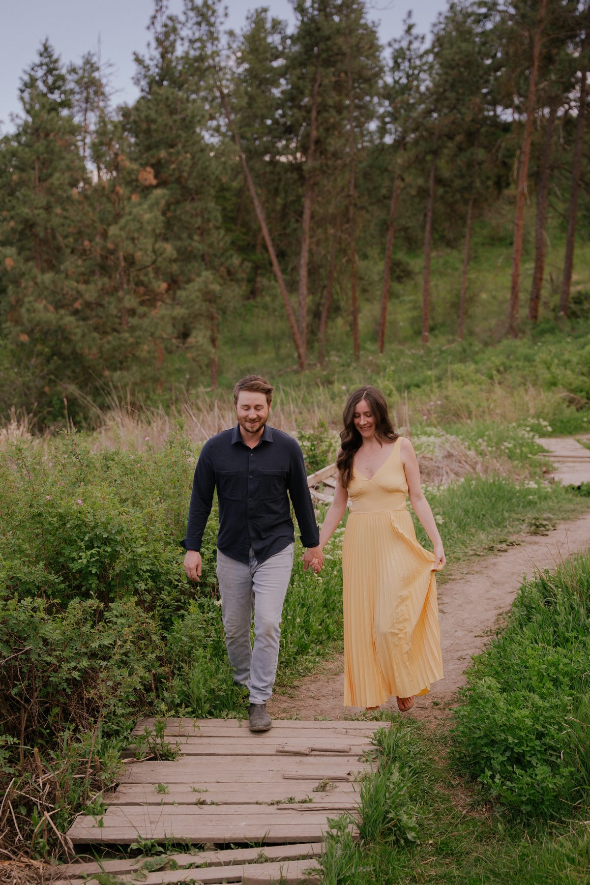 Couple walking hand in hand across a boardwalk on an Okanagan trail, her yellow dress lifting as she goes