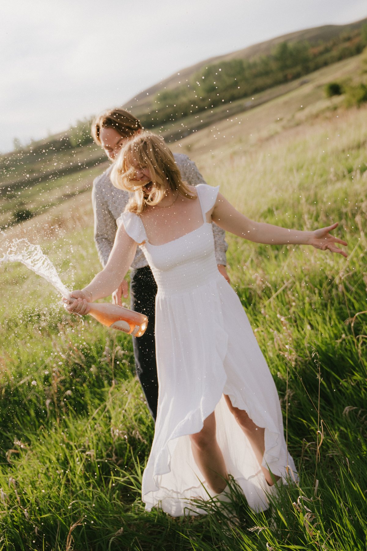 Champagne spraying across the grass as a couple celebrate their engagement on an Okanagan hillside at golden hour