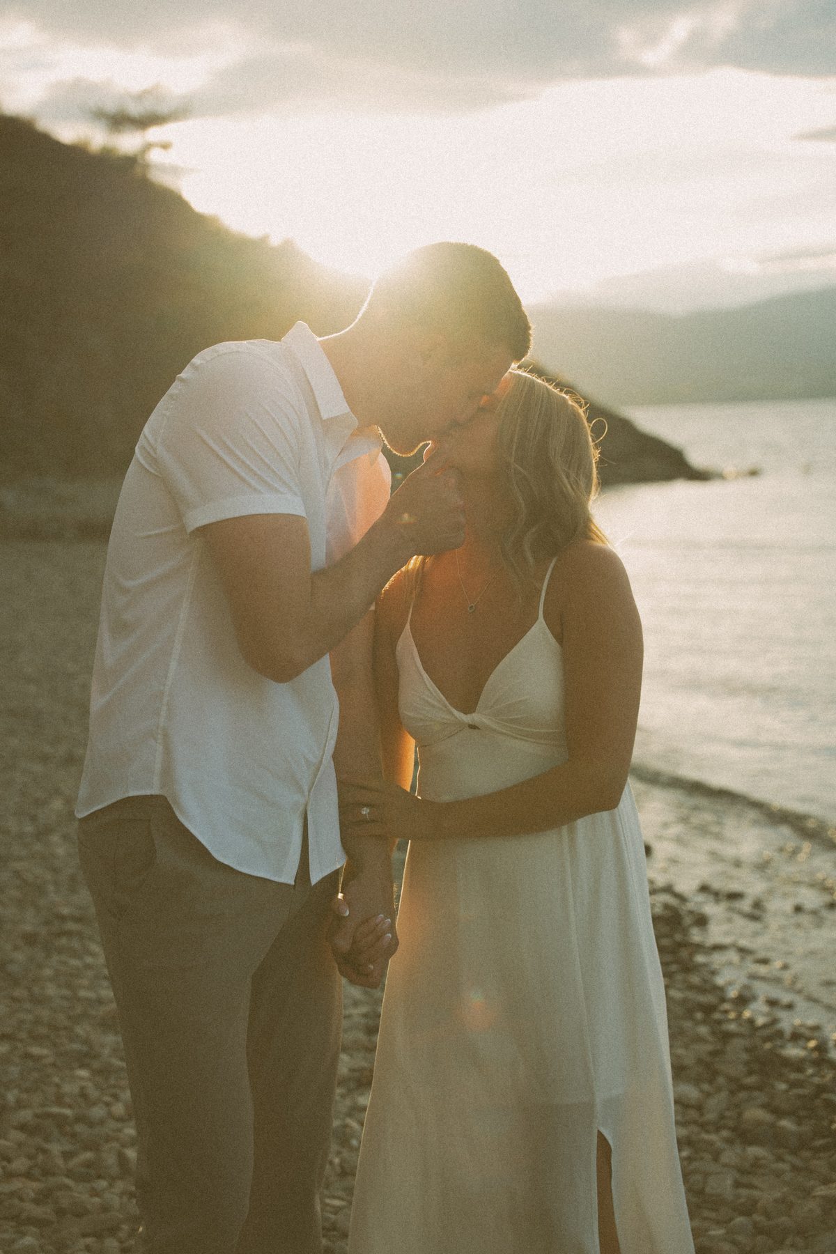He tips her chin up for a kiss on the beach at golden hour, the hills across Okanagan Lake behind them