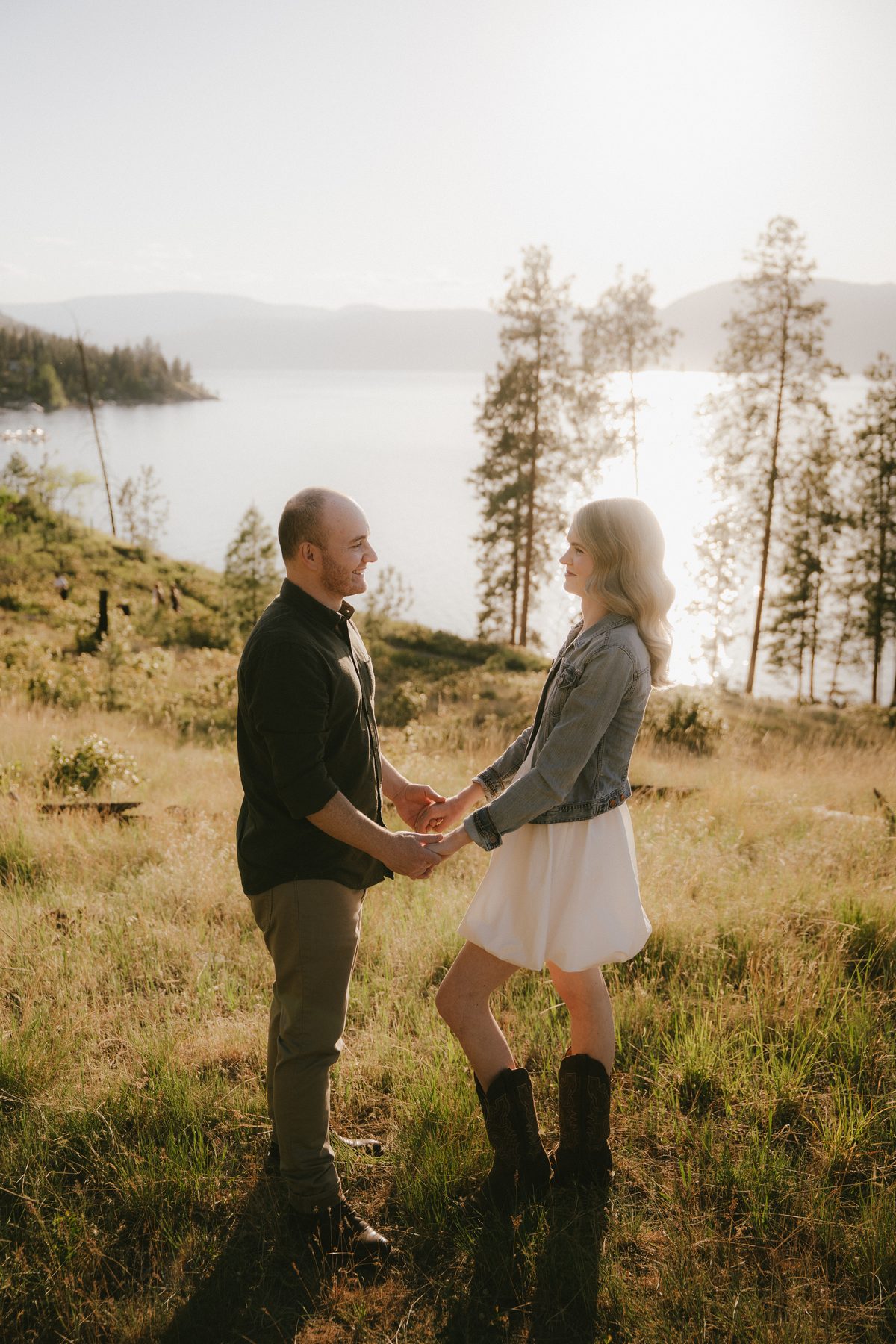 Couple holding hands on a grassy bluff above Okanagan Lake, evening sun on the water below