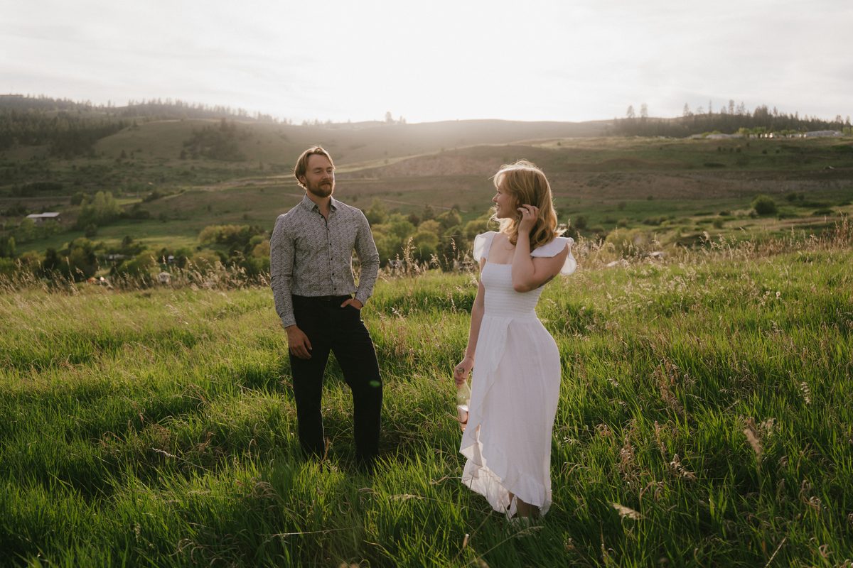 Engaged couple standing apart in a green spring meadow above the Okanagan Valley, the sun low over the hills