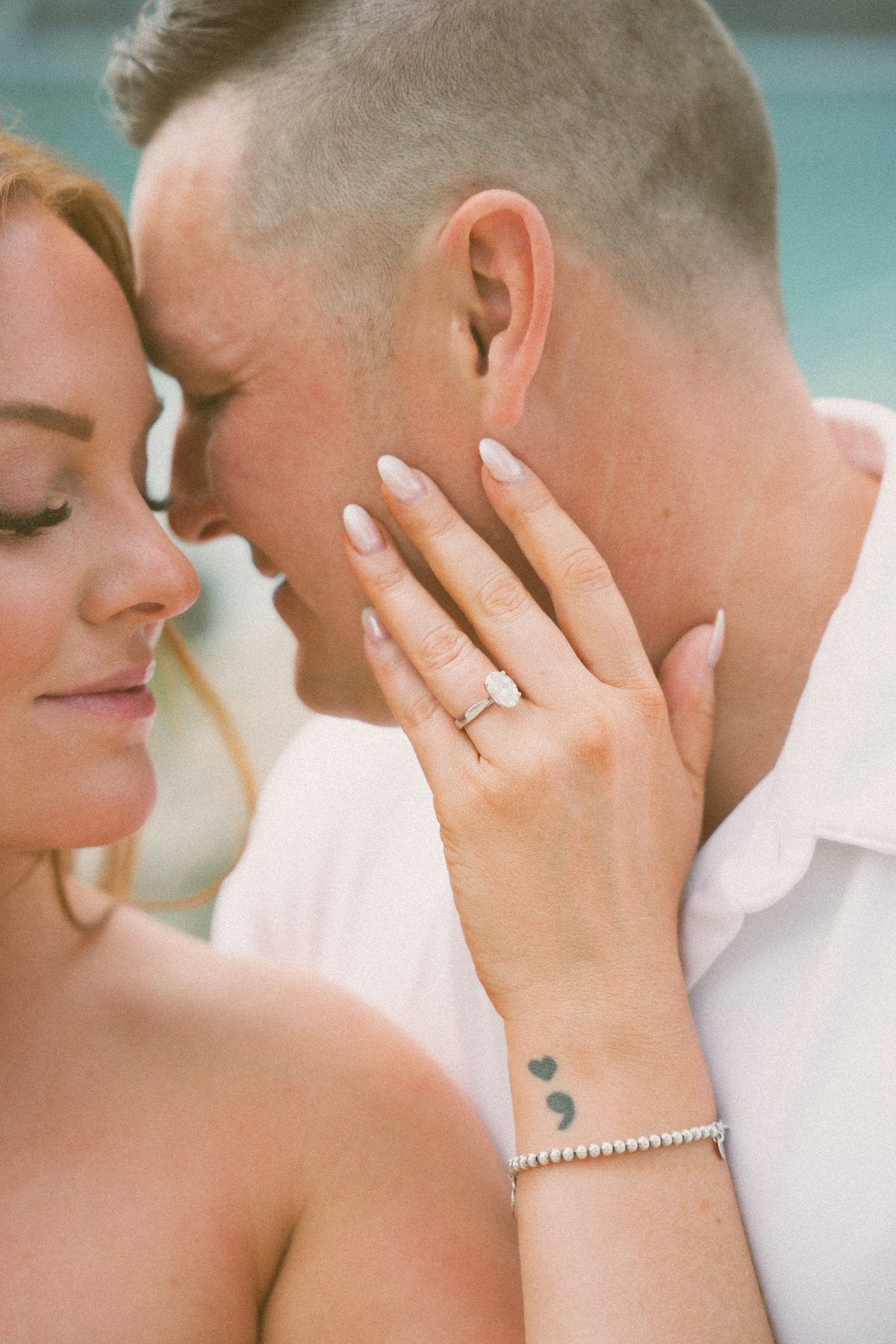 Close-up of her hand resting on his cheek, an oval engagement ring on her finger and a small heart tattoo at her wrist
