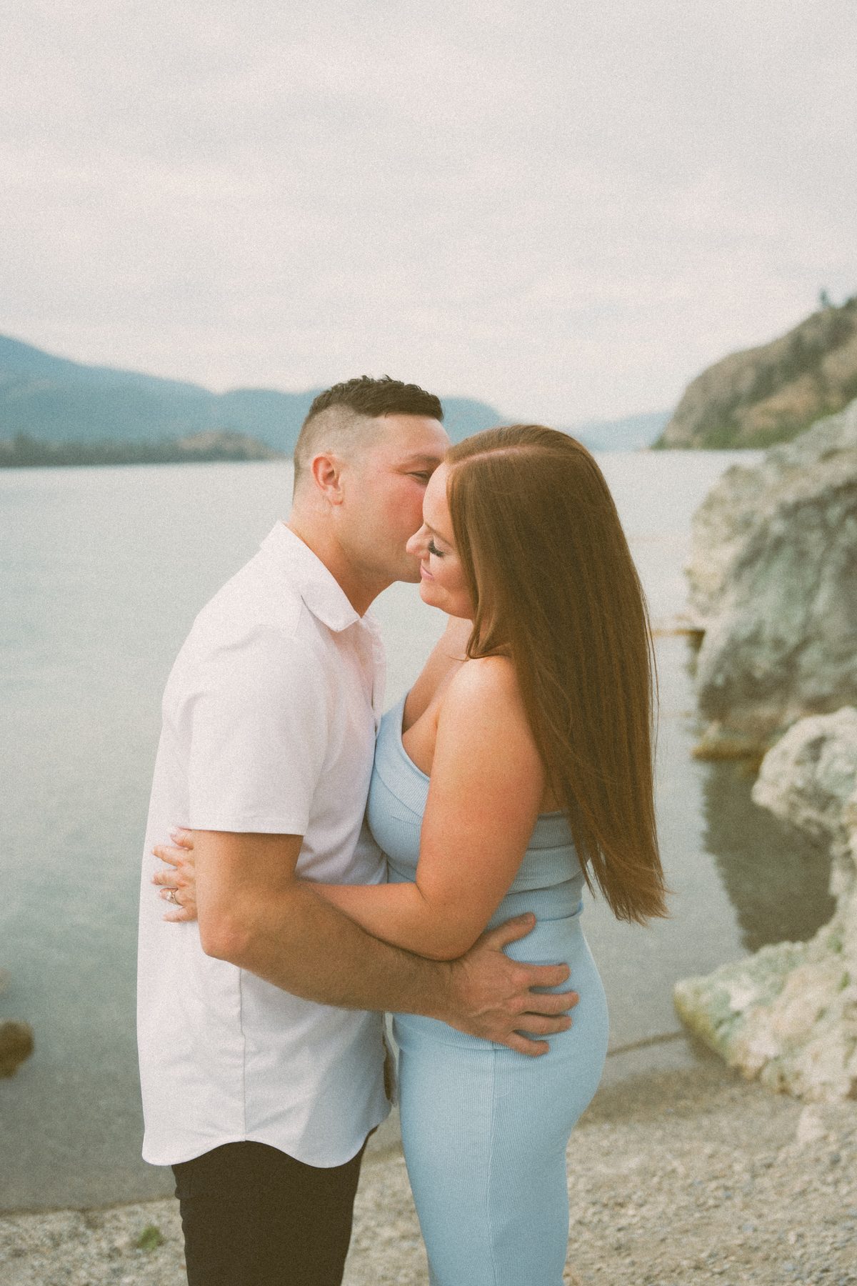 He leans in to kiss her at the water's edge, his arms around her waist on the rocky shore