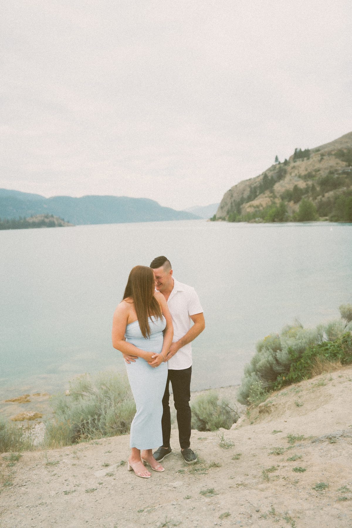 Couple laughing forehead to forehead on a sandy bluff above Okanagan Lake, sagebrush and blue hills around them