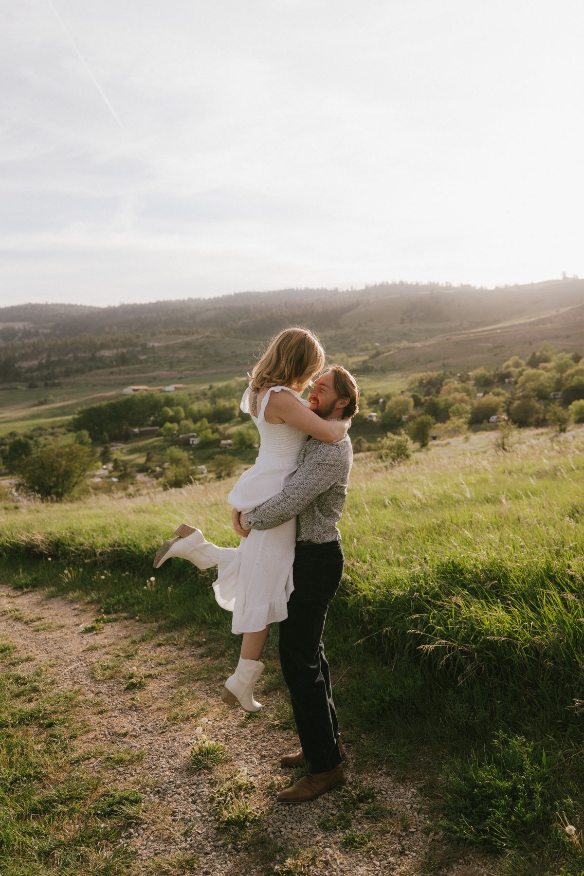 He lifts her off her feet on a hillside track above the Okanagan Valley, evening sun across the benchland behind them