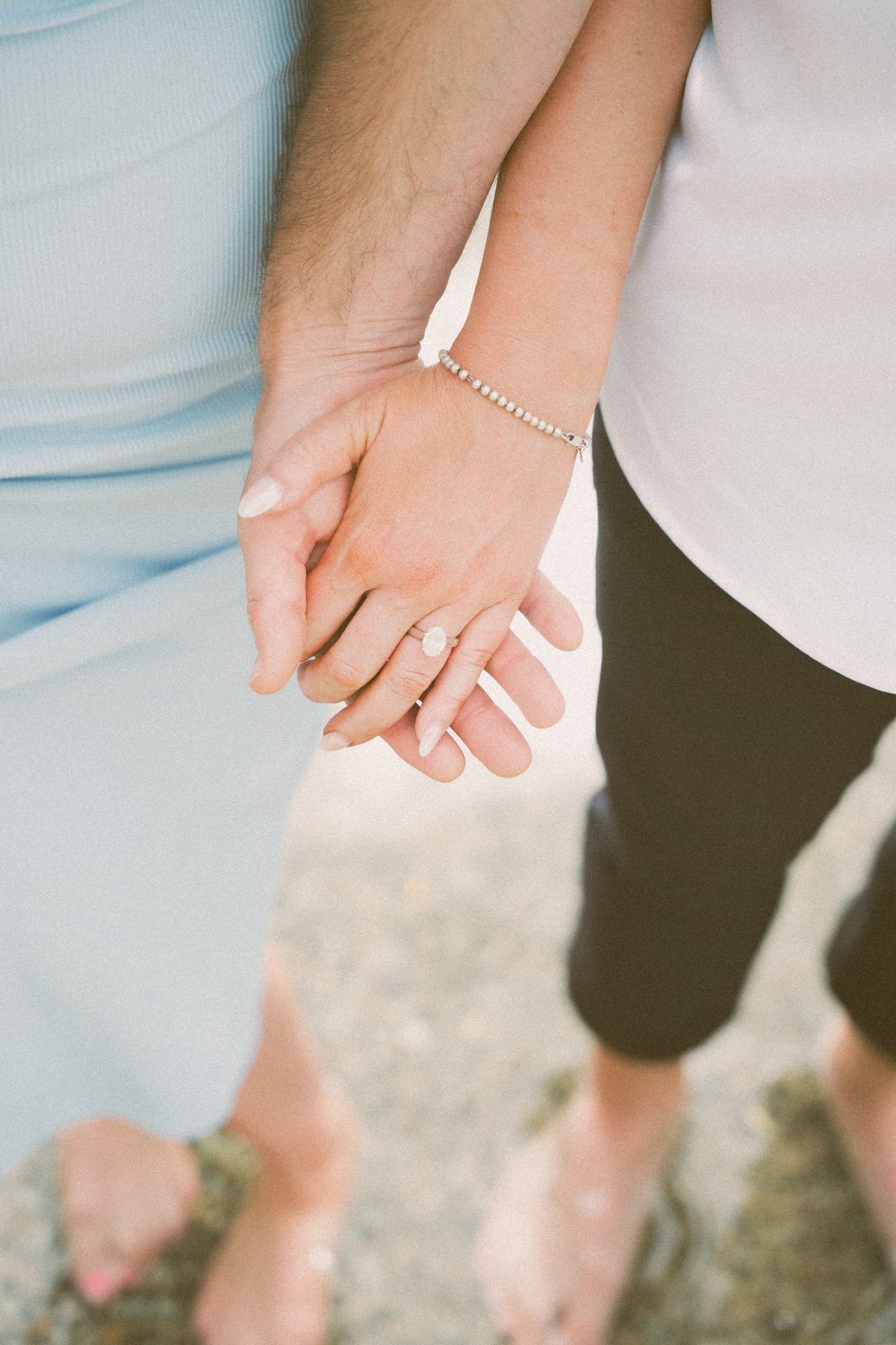 Their hands linked above the shallows, her engagement ring catching the flat afternoon light