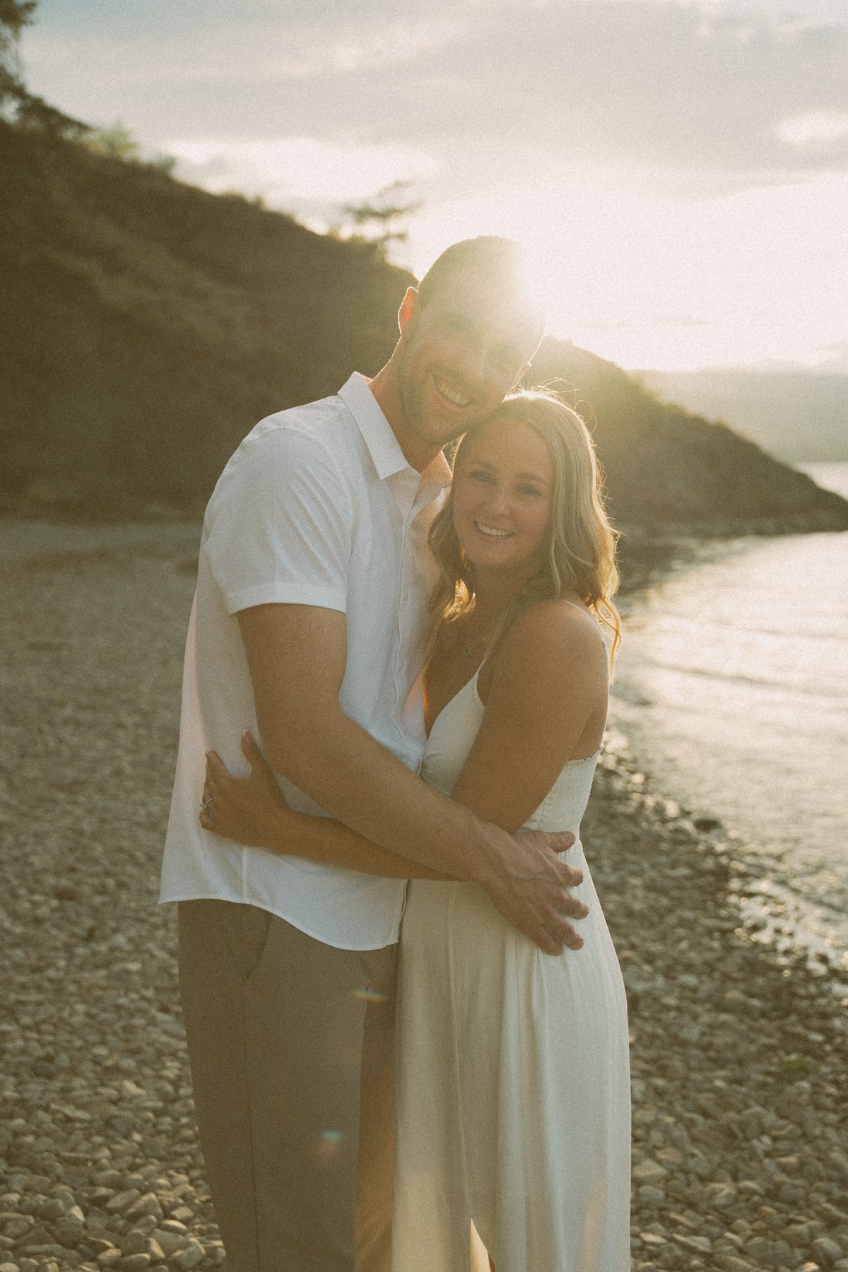 Engaged couple on a pebble beach beside Okanagan Lake, his arms around her from behind, both of them backlit by the evening sun