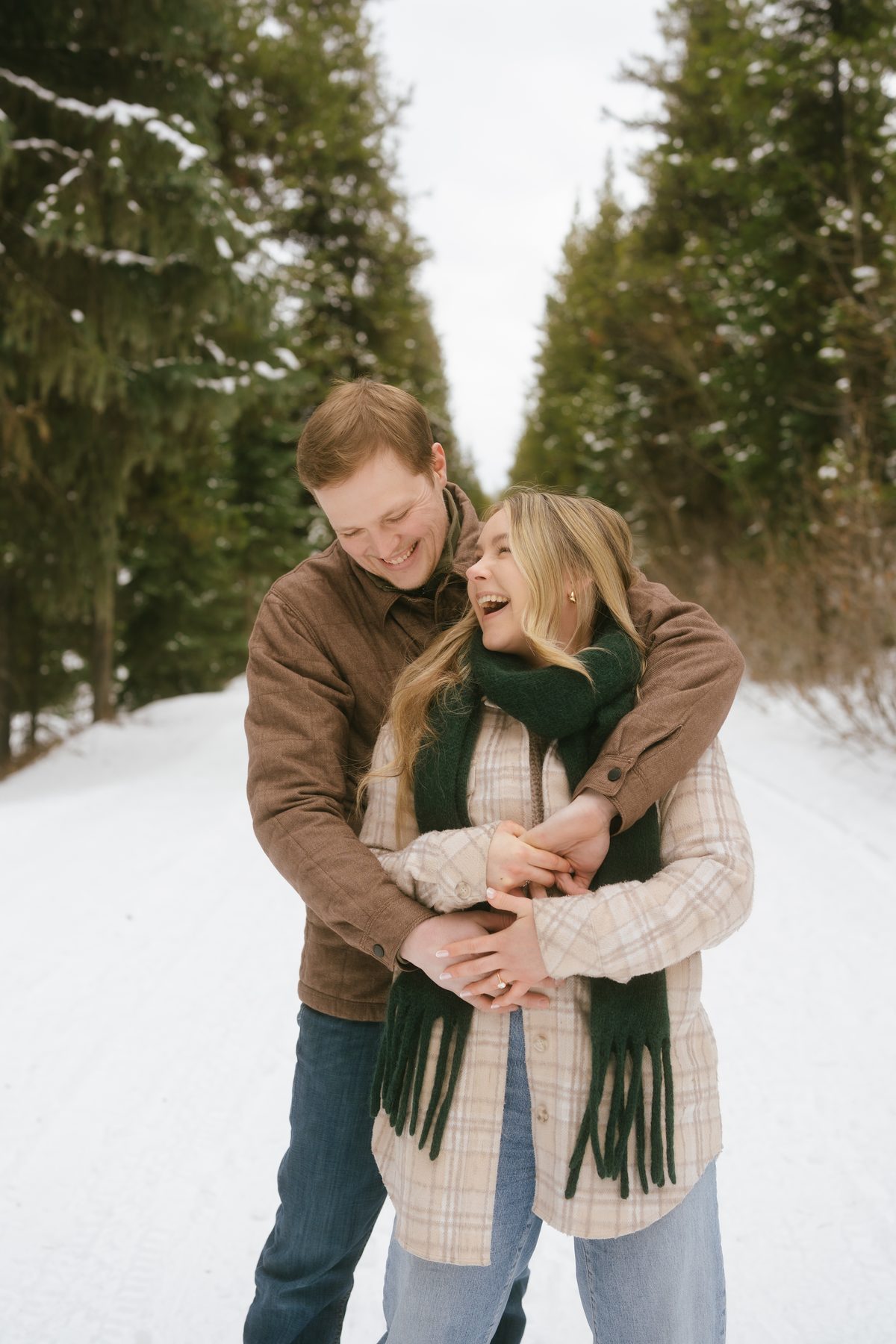 He wraps his arms around her on a snowy forest road, both of them laughing, her engagement ring catching the light