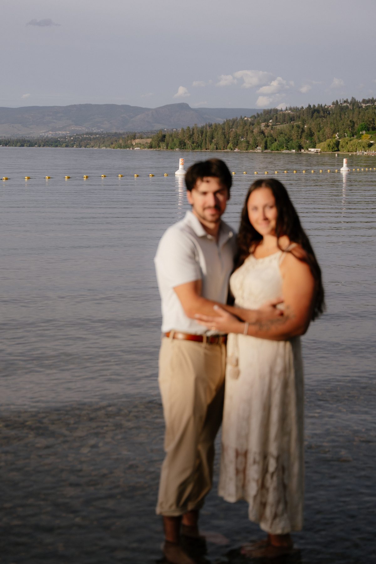 Soft-focus portrait of a couple standing in the shallows of Okanagan Lake, the far shore beyond