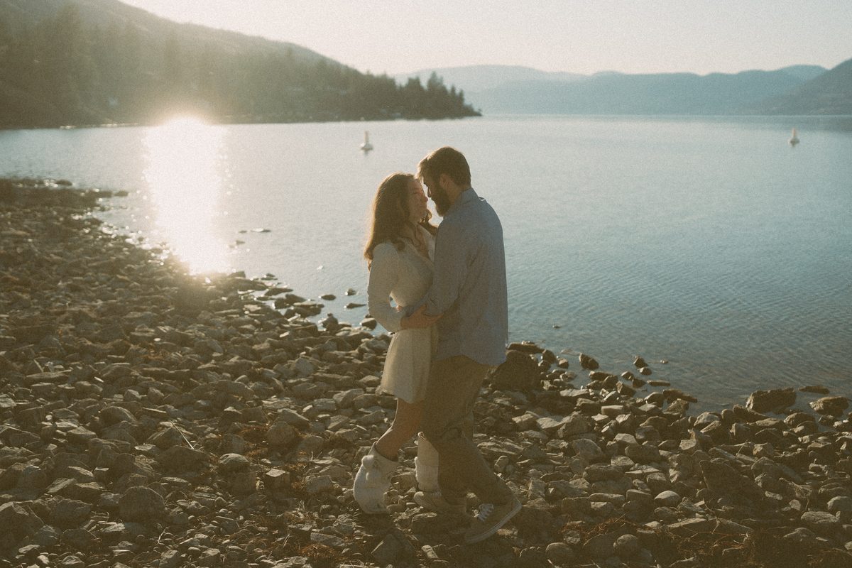 Couple walking the stony shoreline of Okanagan Lake at golden hour, the hills folding away into the distance