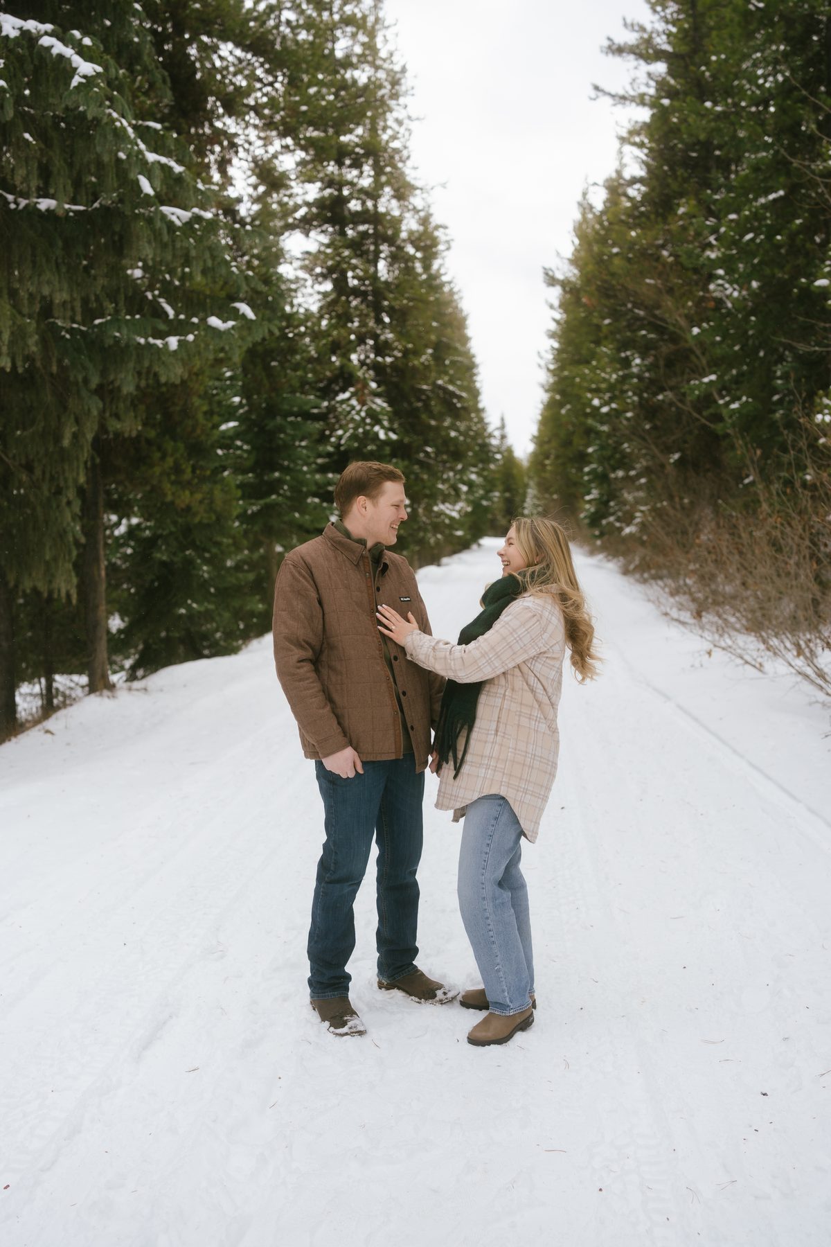Couple laughing on a snow-covered road between rows of evergreens during a winter engagement session