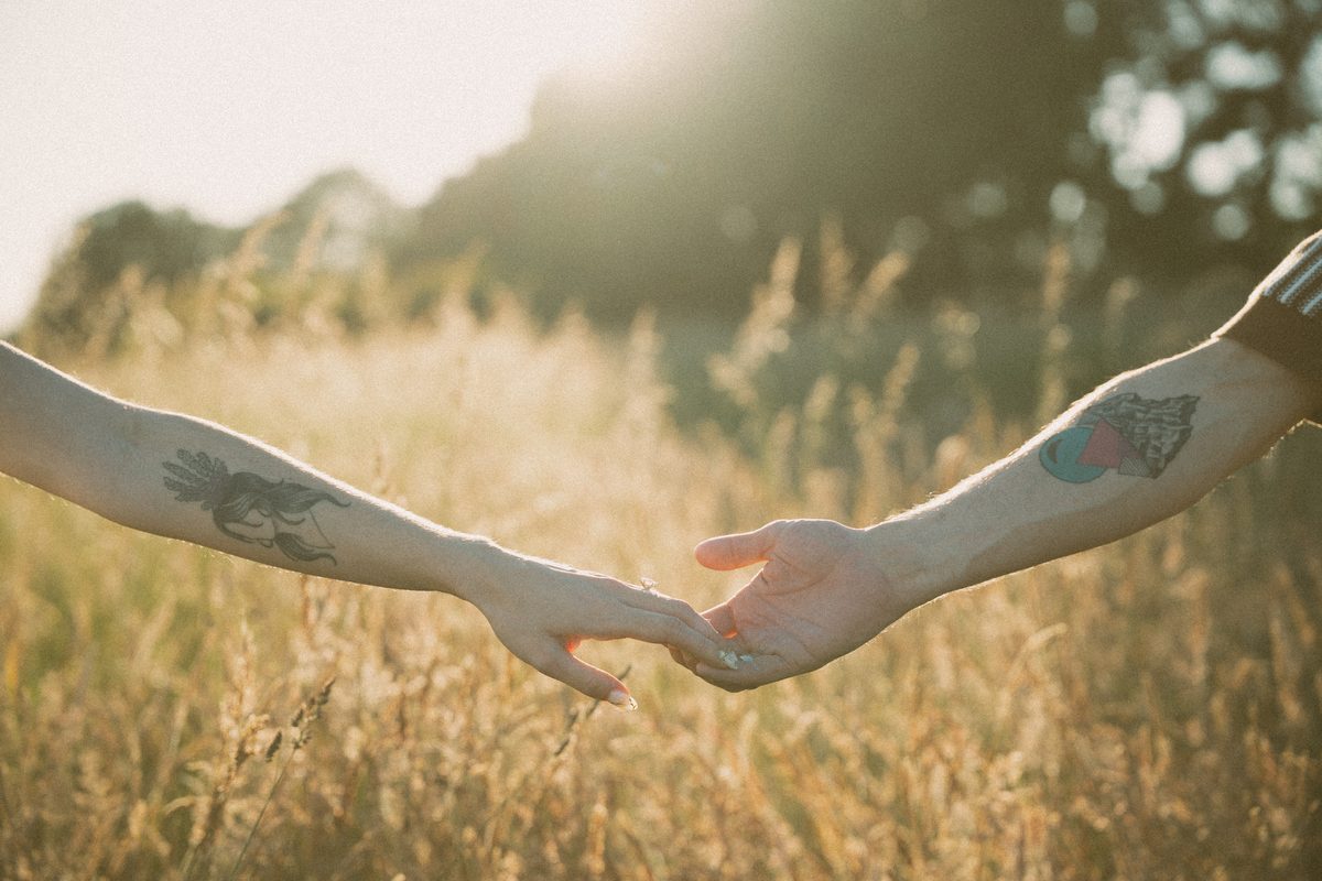 Two hands reaching for each other across dry summer grass, an engagement ring catching the light