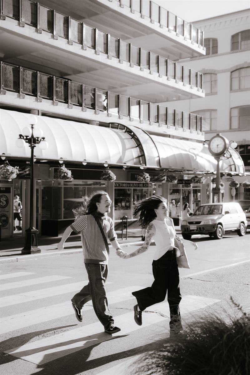 Black and white photo of a couple running hand in hand across a downtown Victoria crosswalk, storefronts and a street clock behind them