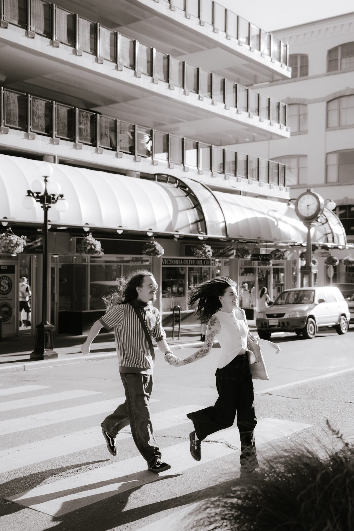 Black and white photo of a couple running hand in hand across a downtown Victoria crosswalk, storefronts and a street clock behind them