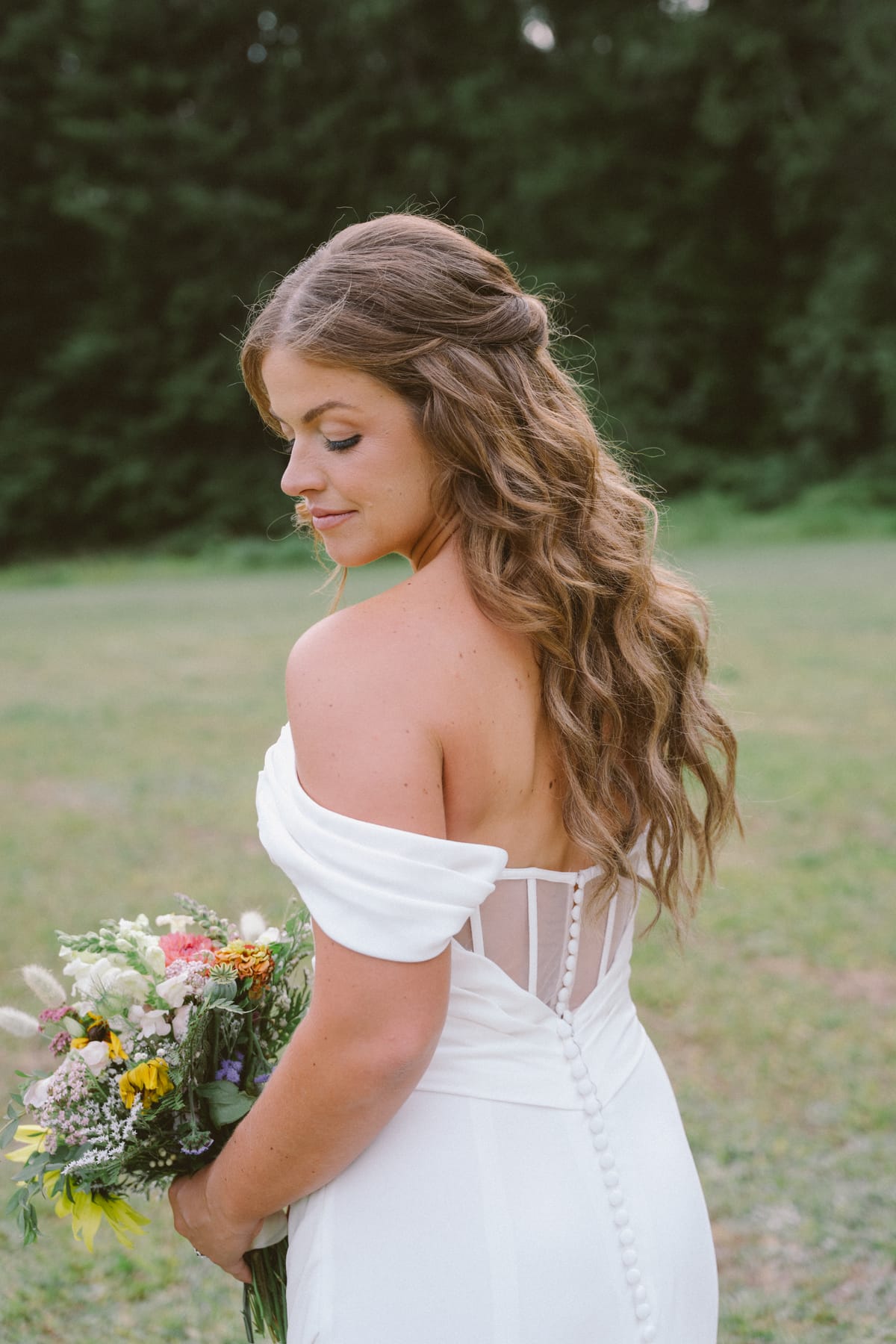 The buttoned back of Kendra's off-shoulder gown as she looks down at her bouquet