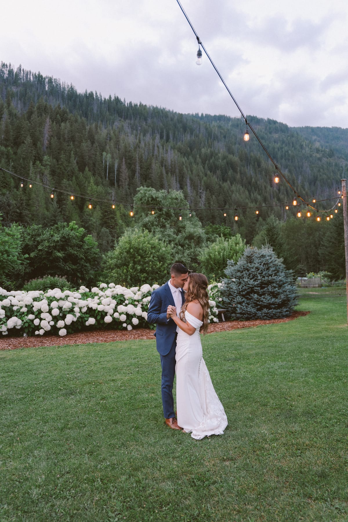 Kendra and Rory dancing by the white hydrangeas under string lights at dusk