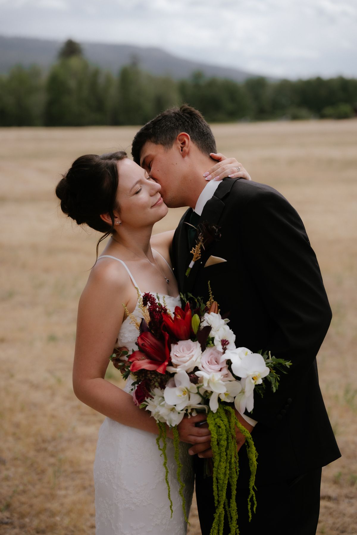 Groom kissing the bride's forehead as she holds her red and white bouquet