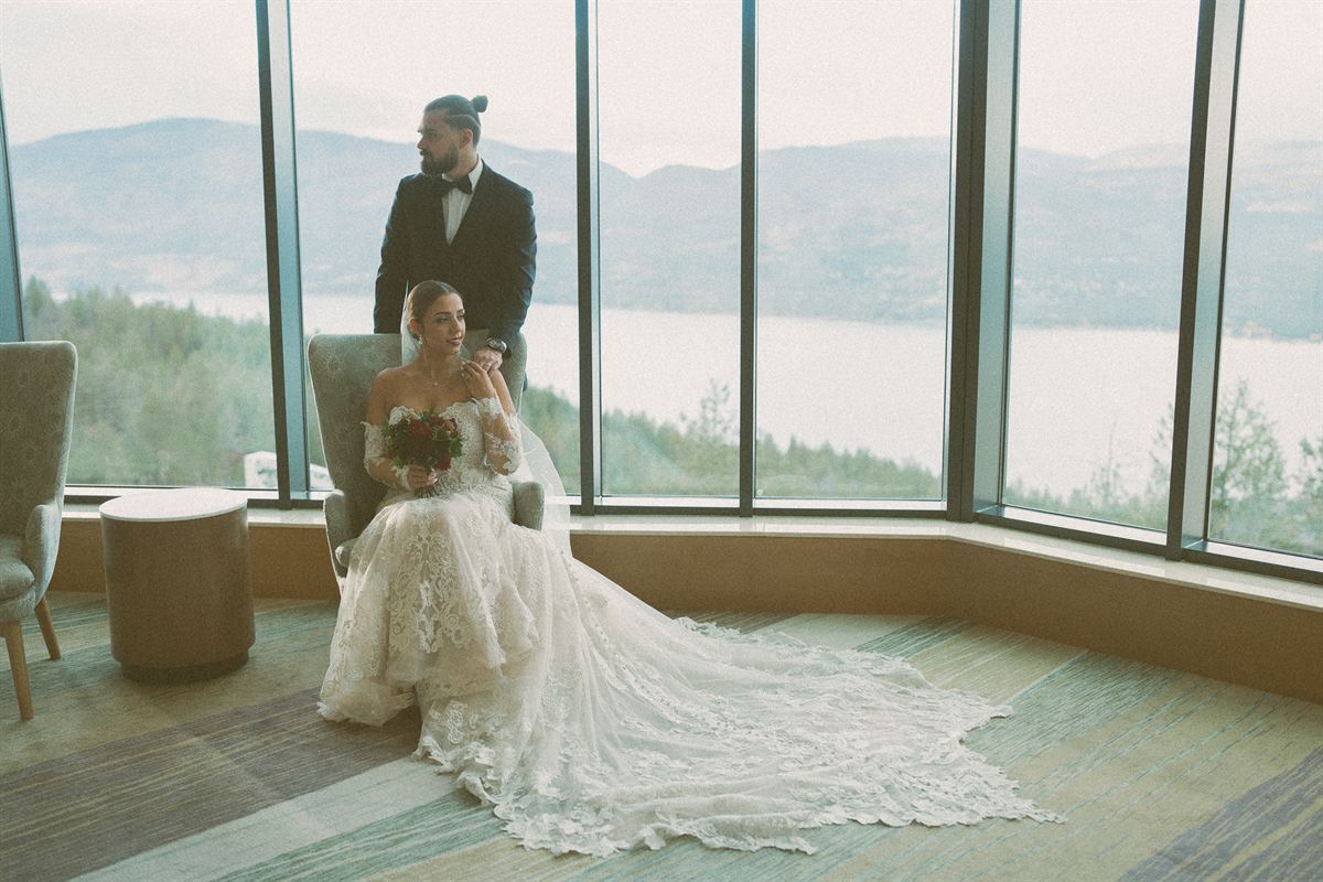 Bride seated in an armchair with her lace train fanned across the floor while the groom stands behind her, floor-to-ceiling windows framing Okanagan Lake and the hills beyond