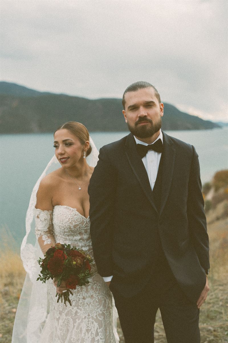 Bride and groom standing together on a bluff above Okanagan Lake under heavy autumn cloud, a deep red bouquet at her side