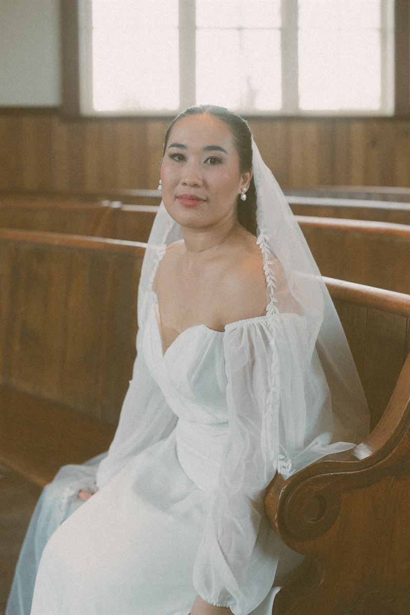 Bride seated in a church pew in a puff-sleeved gown and veil, lit by a tall window