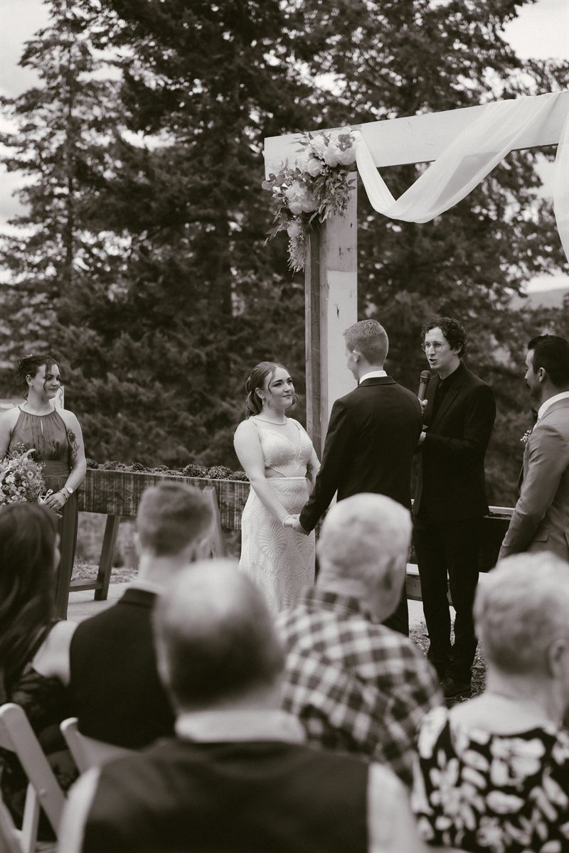 Black and white view from the back of the aisle as the couple face each other during the ceremony, guests watching from folding chairs