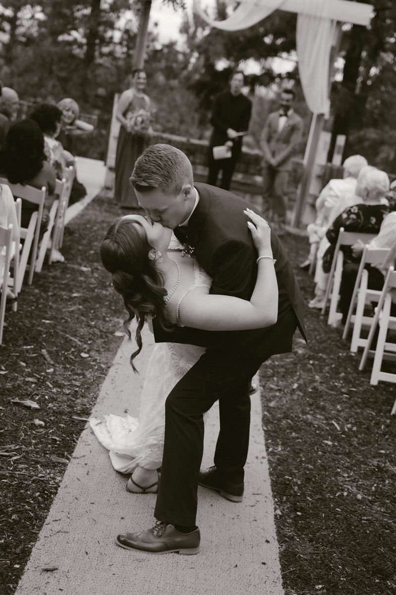 Warm-toned black and white photo of the groom dipping his bride for a kiss in the aisle as their guests applaud