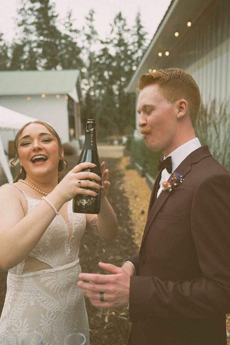 Bride laughing as she and the groom open a bottle of sparkling wine outside the reception barn
