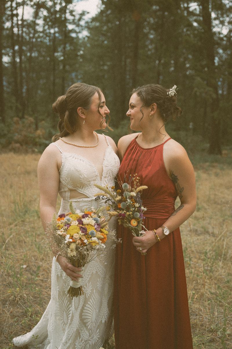 Bride and her bridesmaid smiling at each other in a pine clearing, dried wildflower bouquets in hand