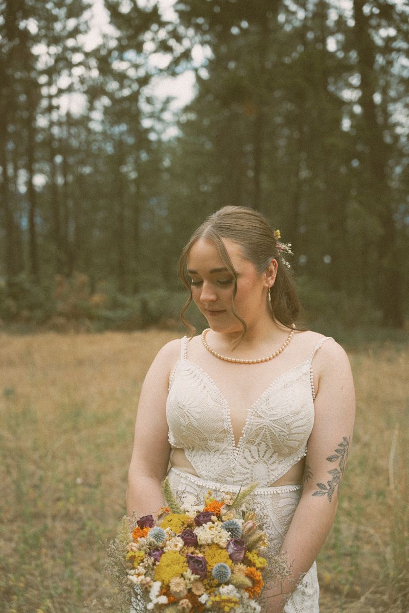Bride looking down at her dried wildflower bouquet in a sunlit clearing, pearls at her throat