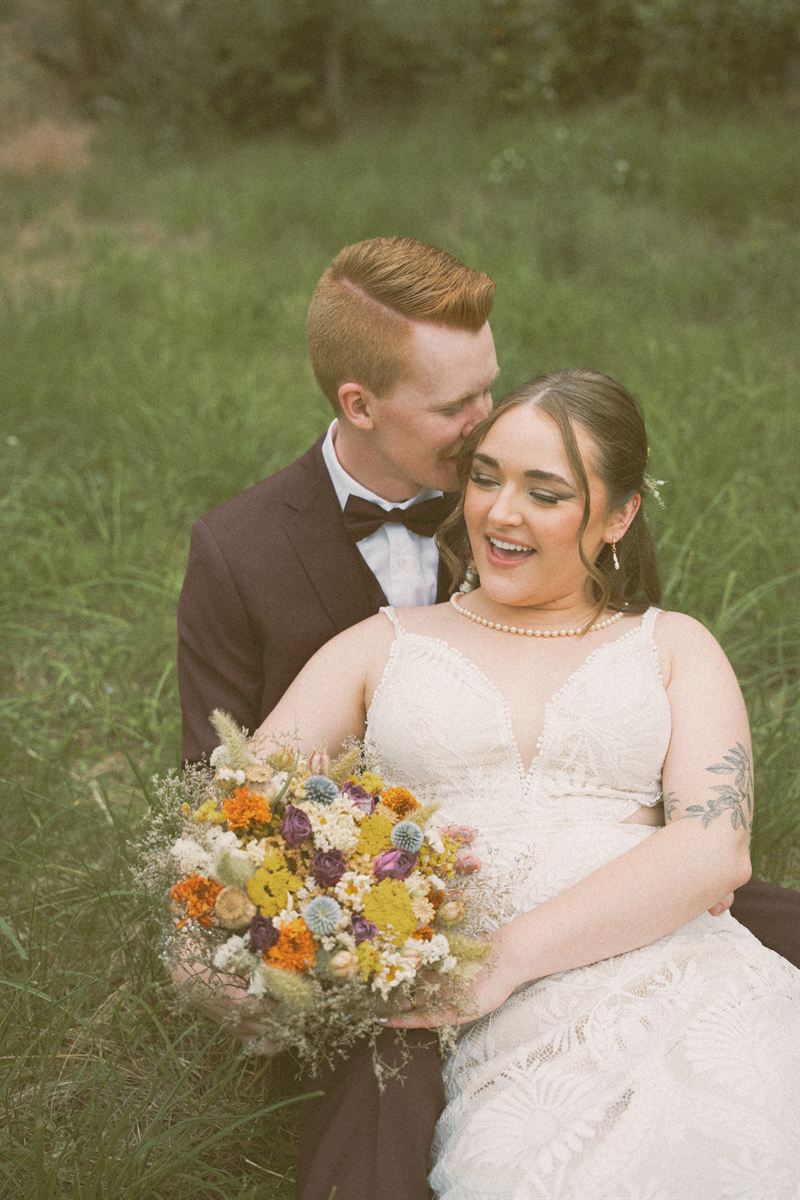 Groom kissing the bride's cheek as she laughs, the two of them sitting in a summer meadow