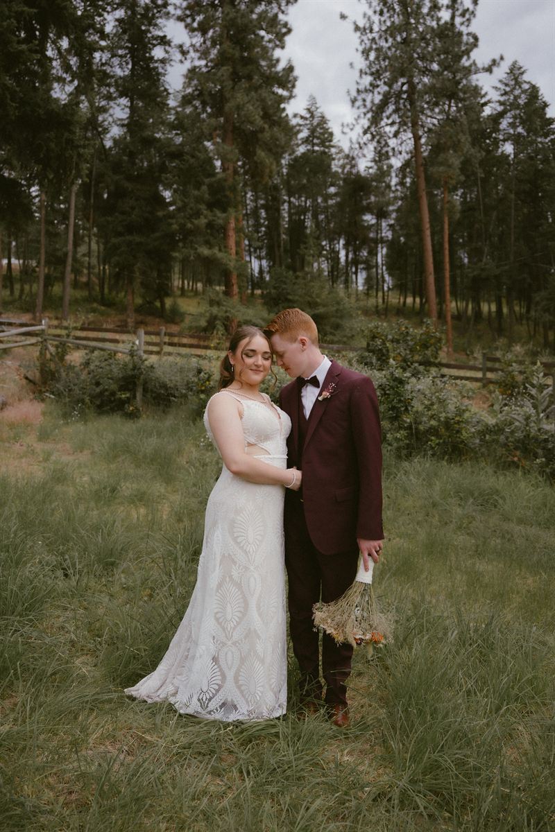 Bride and groom stand forehead to forehead in a meadow, a split-rail fence and pine forest behind them