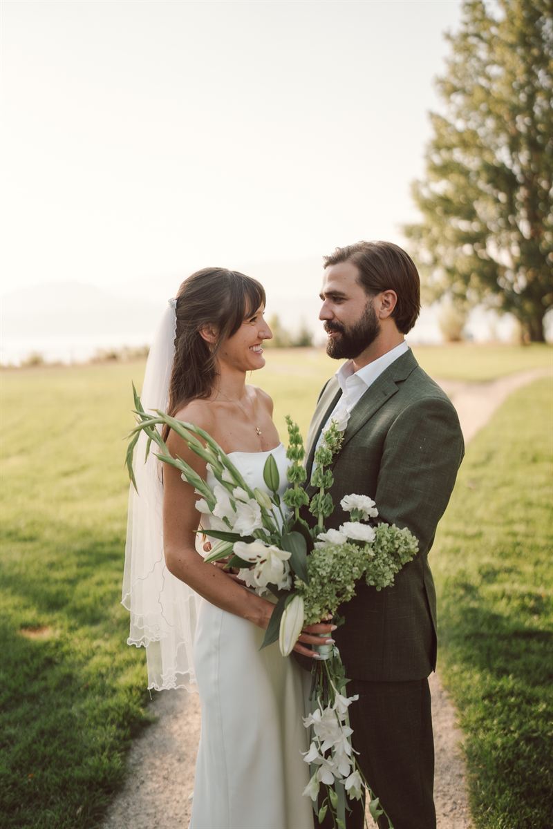 Bride and groom smiling at each other beside Okanagan Lake in Kelowna at golden hour, a white gladiolus bouquet between them