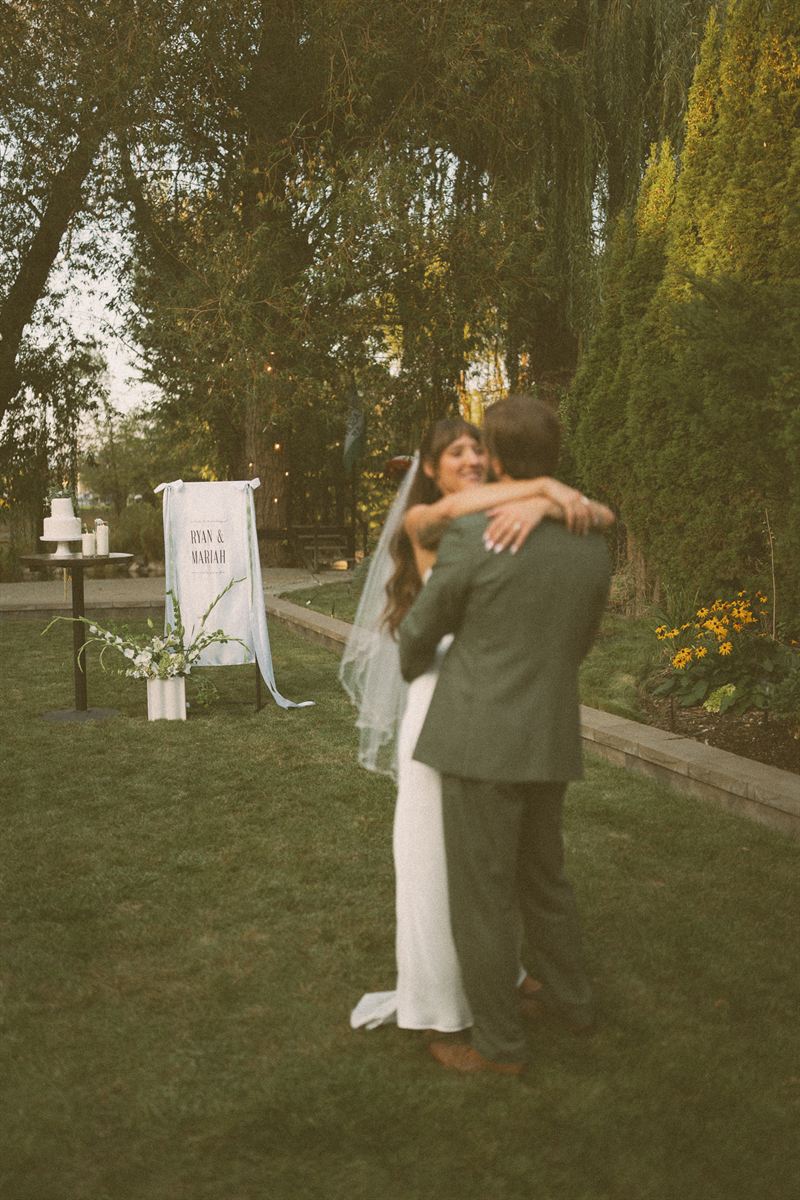 Bride and groom holding each other on the lawn beneath a willow at their Kelowna reception, their names on a sign beside the cake table