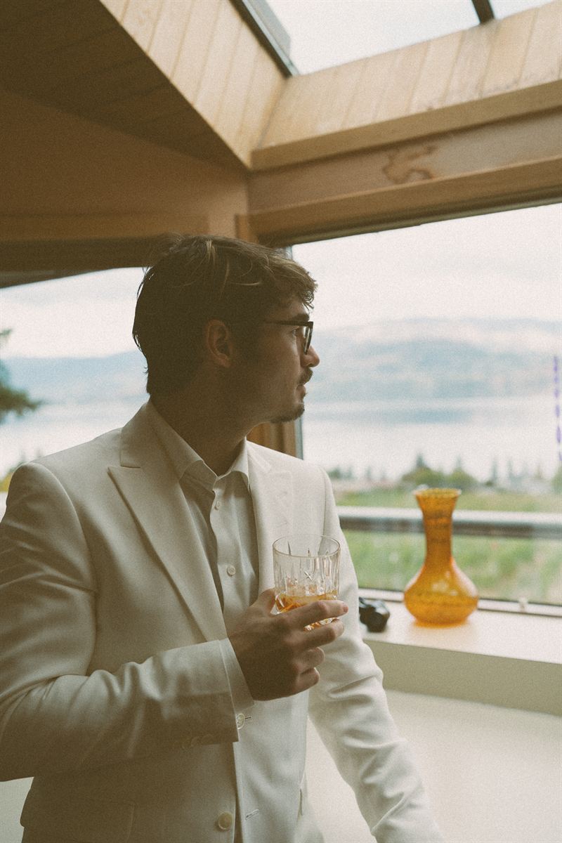 Groom in a cream suit looking out over Okanagan Lake with a glass of whisky before the ceremony