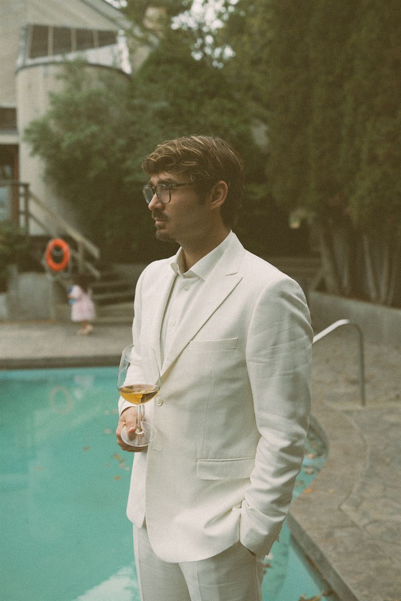 Groom in a cream suit standing beside a pool with a glass of wine, waiting for the day to begin
