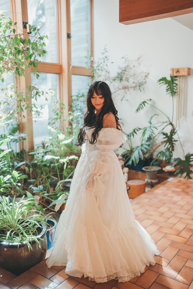 Bride in a tiered vintage gown looking down at her skirt in a plant-filled room with terracotta tile
