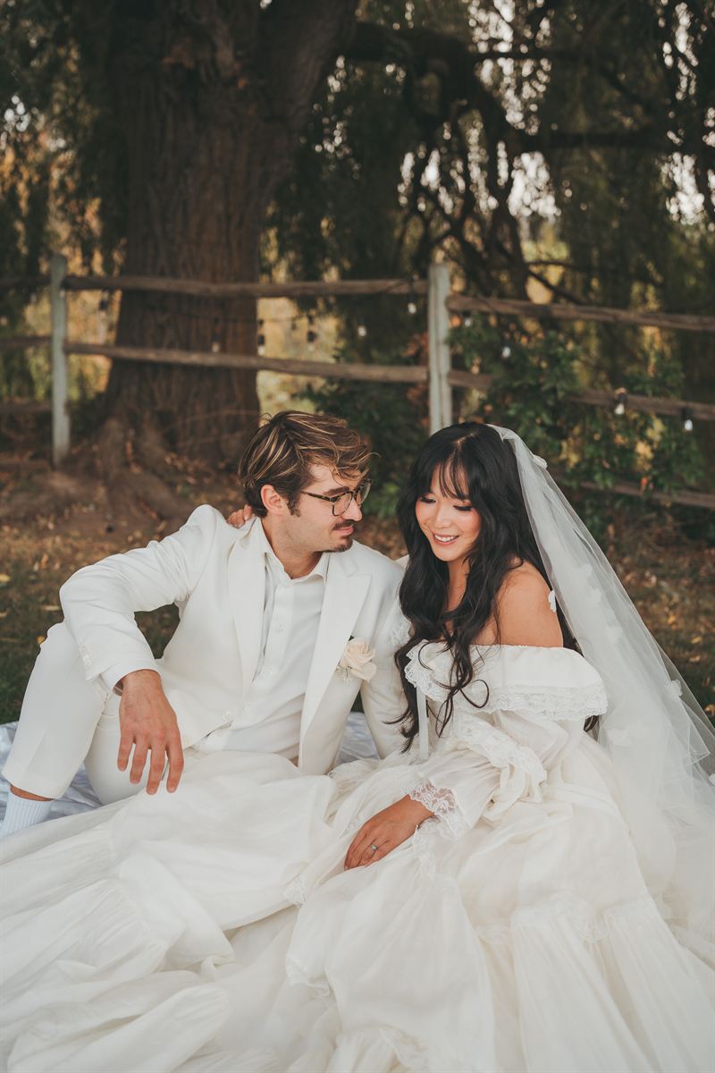 Bride and groom sitting together on a blanket beneath a willow tree, her vintage gown spread around her