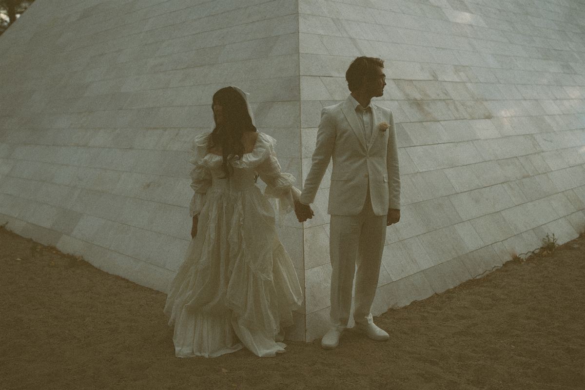 Bride and groom hold hands on either side of a curved stone wall, both dressed in ivory