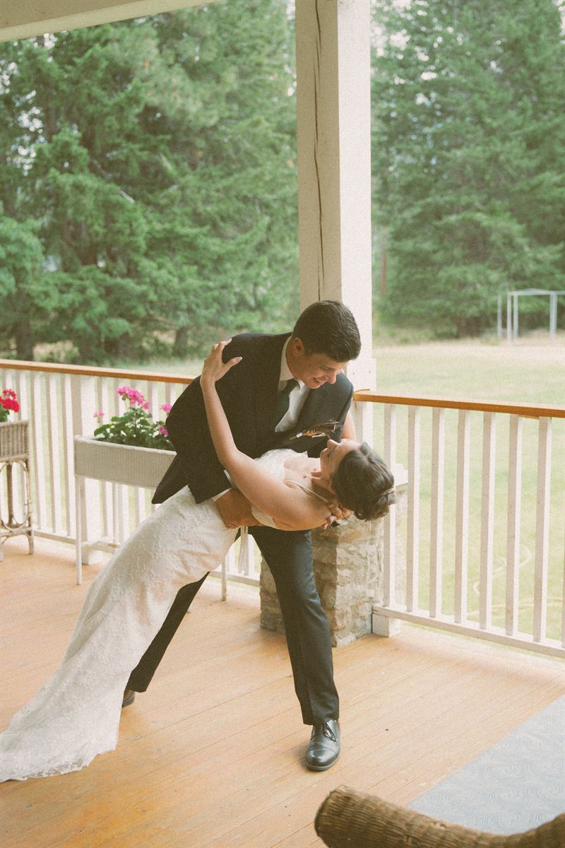 Groom dipping his bride for a kiss on a covered porch, geraniums in the window box and forest beyond