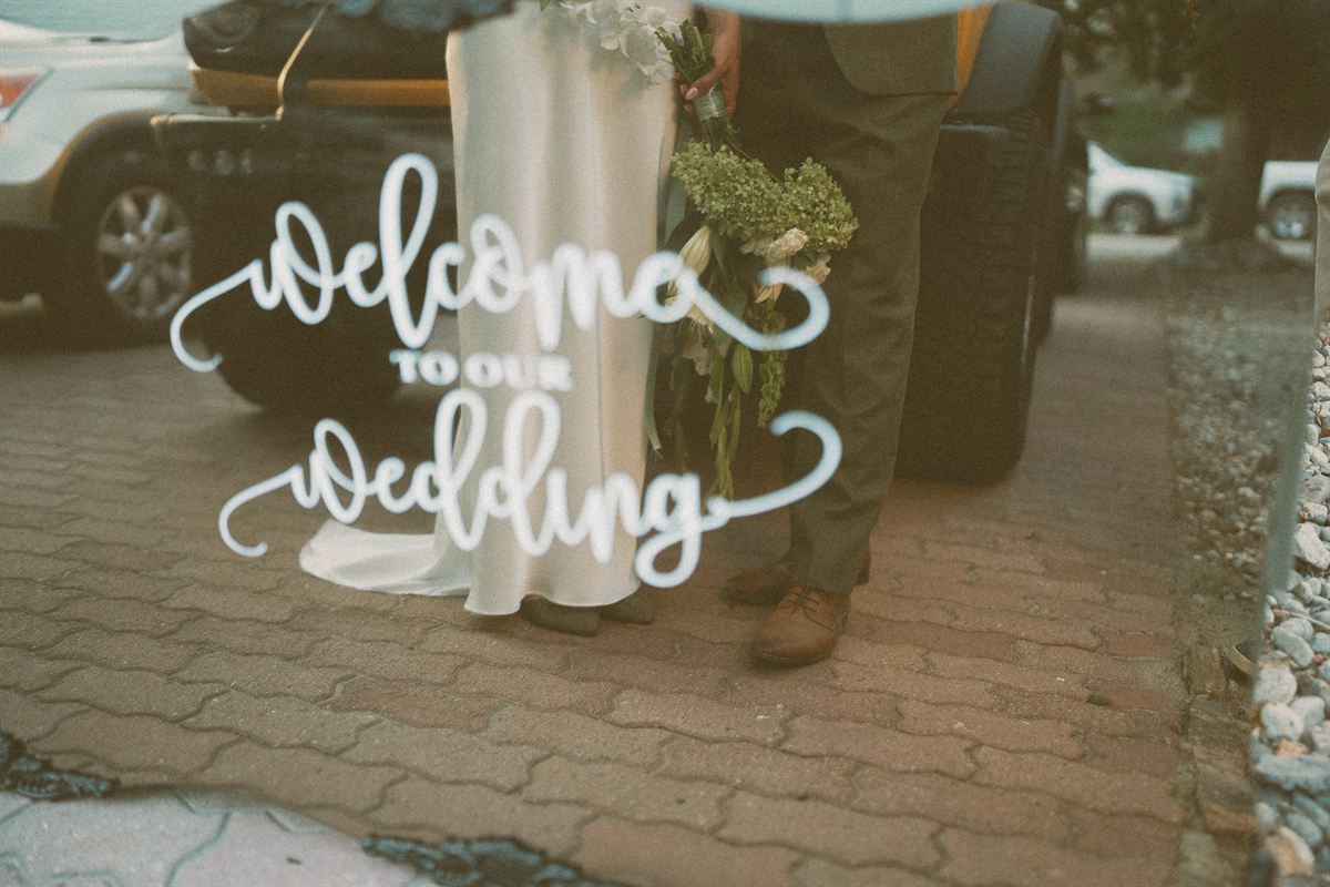 A hand-lettered welcome sign carried past the parked cars at a Kelowna wedding
