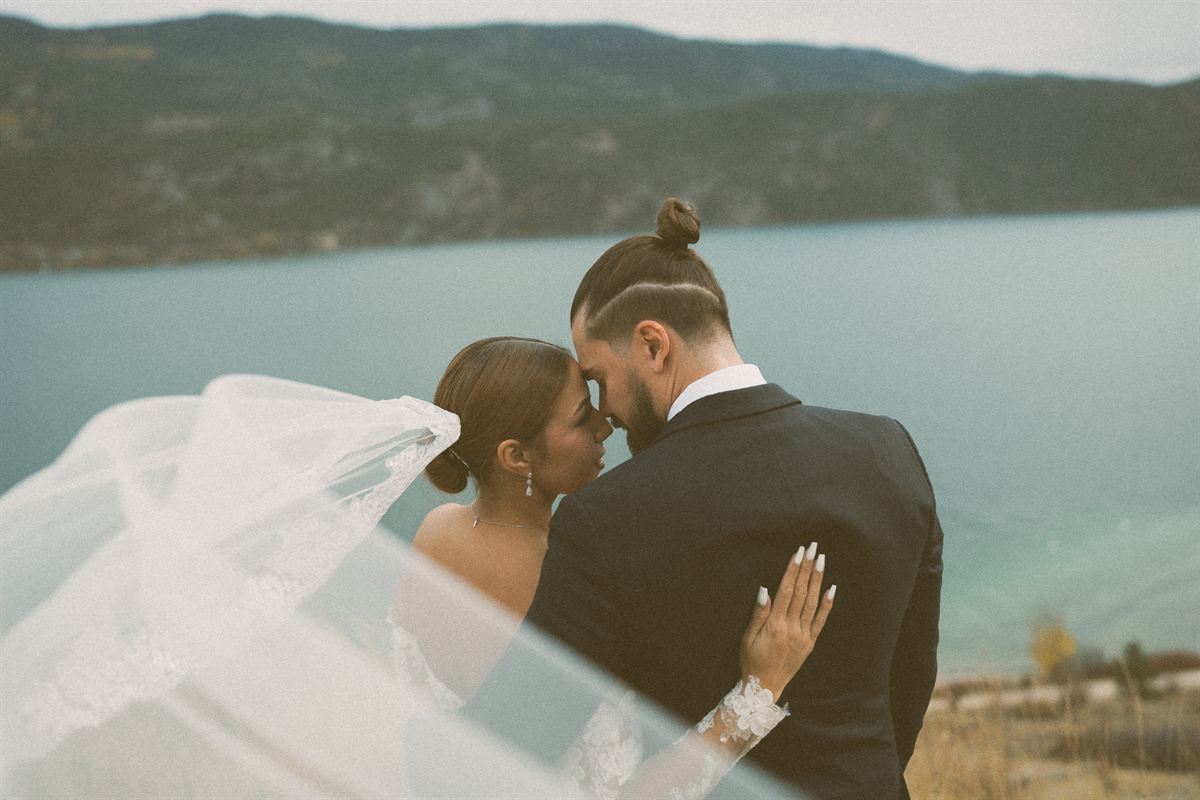 The bride's veil sweeps across the frame on the wind as she and the groom touch foreheads above the turquoise water of Okanagan Lake